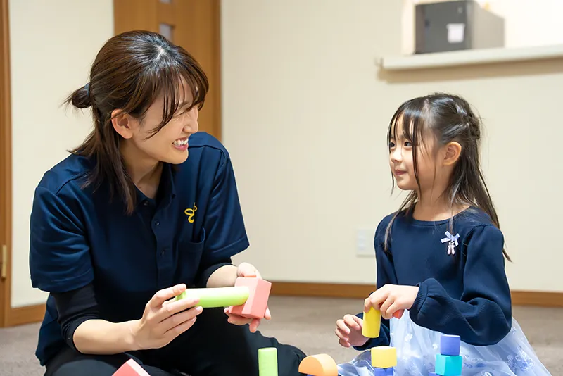 スタッフが子どもと笑顔で向き合い、カラフルな積み木を使って一緒に遊んでいる様子